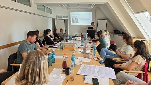 Personen sitzen an einem langen Tisch in einem hellen Raum und schauen auf eine Präsentation an der Wand
