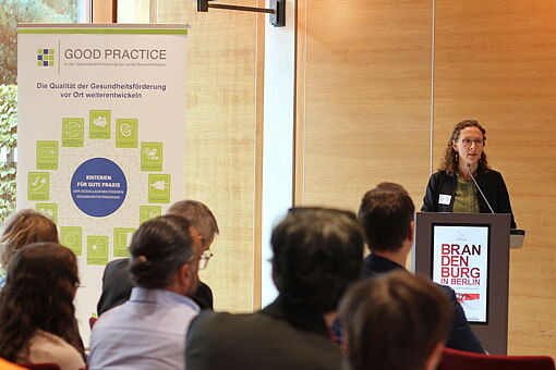 Frau am Rednerpult mit Schild 'BRANDENBURG IN BERLIN' spricht vor sitzendem Publikum, links Roll-up mit Text 'GOOD PRACTICE Die Qualität der Gesundheitsförderung vor Ort weiterentwickeln'.