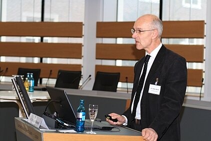 8.	Foto zeigt Dr. Volker Wanek beim Vortrag am Rednerpult, auf dem sich ein Laptop befindet