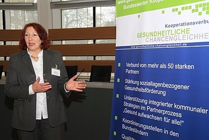 10.	Foto zeigt Frau Prof. Dr. Elisabeth Pott neben einem Rollup zum Kooperationsverbund Gesundheitliche Chancengleichheit