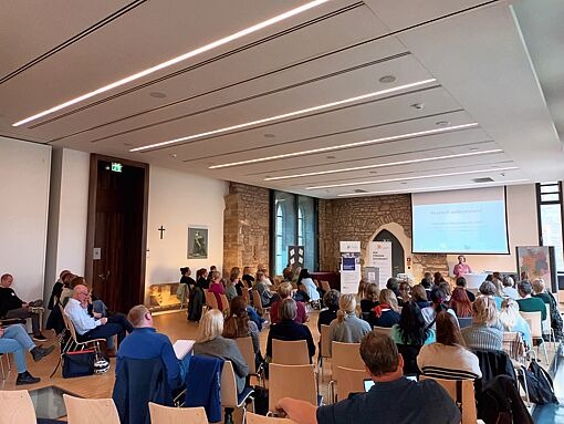 Großer Seminarraum mit vielen sitzenden Personen, Vortragende vor Leinwand mit Präsentation, helle Decke mit Linienbeleuchtung.