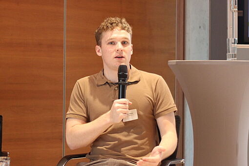 Zwei Männer sitzen auf Stühlen, einer hält ein Mikrofon, beide tragen Namensschilder, im Hintergrund Holzpaneele und ein Stehtisch.