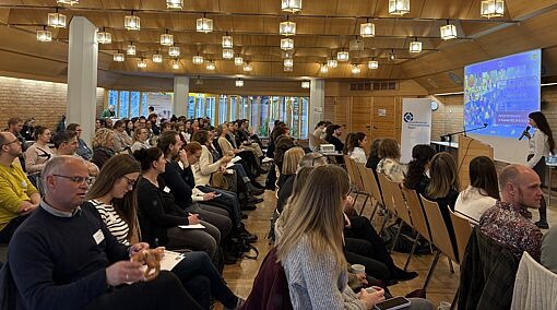 Großer Seminarraum mit vielen sitzenden Teilnehmenden, die einer Präsentation an einem Bildschirm folgen.