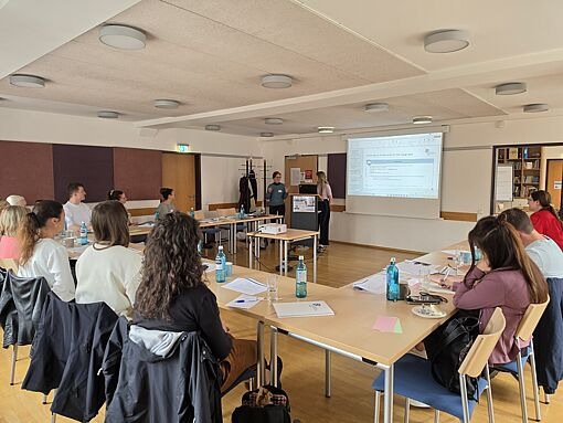Mehrere Personen sitzen an Tischen in U-Form in einem Seminarraum, zwei Personen stehen vorne neben einem Beamerbildschirm mit Präsentation.
