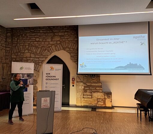 Person steht vor einem Rednerpult und präsentiert eine Folie mit dem Titel 'Einsamkeit im Alter - warum braucht es AGATHE?'.