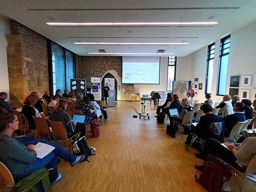 Mehrere Personen sitzen in einem hellen Raum mit Steinwand und großen Fenstern, einige mit Laptops, vor einer Leinwand mit Präsentation.