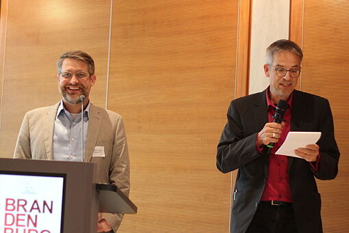 Zwei Männer bei der jährlichen Tagung des Kooperationsverbundes Gesundheitliche Chancengleichheit, einer, Dr. Christoph Aluttis, steht am Rednerpult mit dem Schriftzug 'BRANDENBURG IN BERLIN', der andere, Stefan Bräunling, hält ein Mikrofon und ein Blatt Papier.