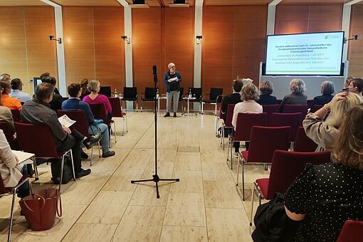 Vortragende steht vor Publikum in einem Konferenzraum mit Holzverkleidung, auf Leinwand Text zum jährlichen Treffen des Kooperationsverbundes Gesundheitliche Chancengleichheit.