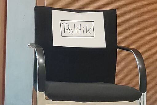 Schwarzer Bürostuhl mit Armlehnen und Rollen, auf der Rückenlehne ein weißes Schild mit der Aufschrift 'Politik', vor einer Holzpaneelwand.