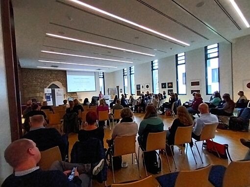 Mehrere Personen sitzen in einem hellen Raum mit hohen Fenstern und sehen auf eine Leinwand mit Präsentation.