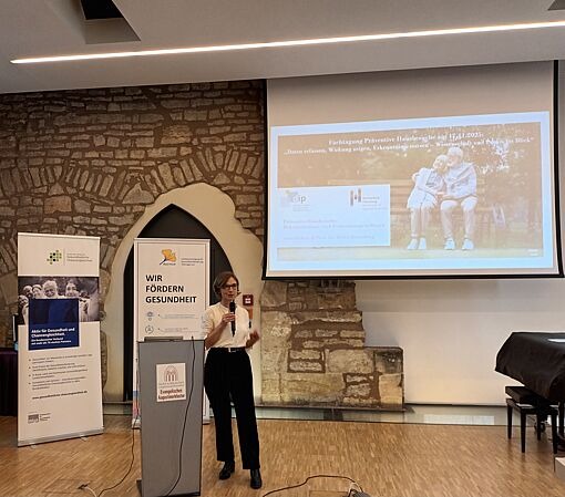 Frau steht mit Mikrofon vor einem Rednerpult in einem Raum mit Steinwand und zwei Roll-ups, rechts ein Flügel, an der Wand eine Projektionsleinwand mit Bild von zwei älteren Menschen auf einer Bank.