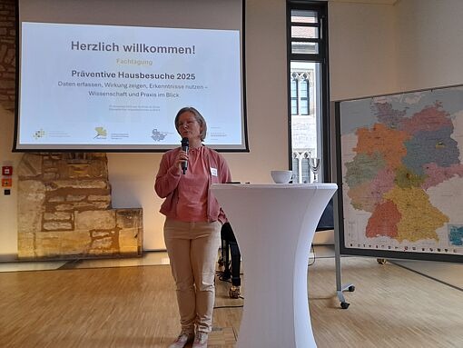 Frau steht mit Mikrofon vor einem Stehtisch in einem Raum mit Projektionsleinwand und Deutschlandkarte an der Wand.