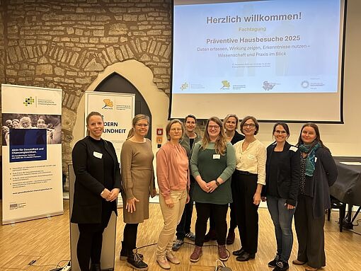 Gruppe von neun Personen steht vor einer Leinwand mit der Überschrift 'Herzlich willkommen! Fachtagung Präventive Hausbesuche 2025'. Im Hintergrund zwei Roll-ups mit Text und Logos.