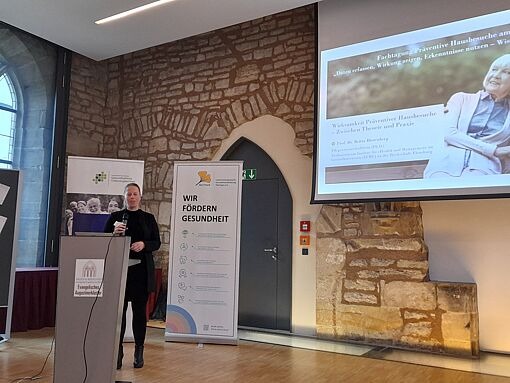 Person steht an einem Rednerpult in einem Raum mit Steinmauer, neben zwei aufgestellten Bannern und einer Projektion an der Wand.