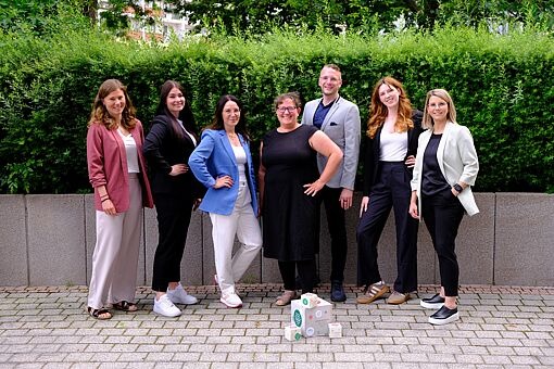 Sieben Personen in Business-Kleidung stehen vor einer Hecke auf gepflastertem Boden und posieren mit einem Stapel aus weißen Kartons auf dem Boden