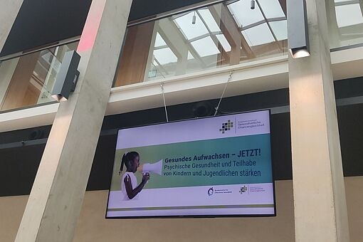 Großer Bildschirm in modernem Gebäude zeigt Präsentation mit Titel 'Gesundes Aufwachsen – JETZT! Psychische Gesundheit und Teilhabe von Kindern und Jugendlichen stärken'.